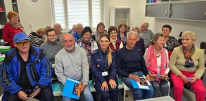 Grupa seniorów siedzących na ławkach, po środku umundurowana policjantka.