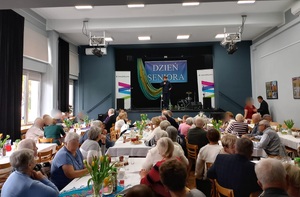 Seniorzy siedzący przy stolikach, na scenie policjantka,  w tle baner z napisem Dzień Seniora
