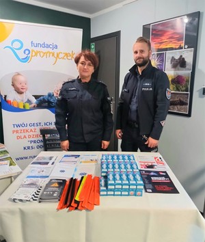 Umundurowana policjantka i policjant, przed nimi na stoliku poukładane ulotki i odblaski.
