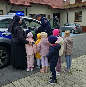 Na zdjęciu grupa przedszkolaków i siostra zakonna oglądający radiowóz, który pokazuje im policjant.