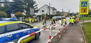 Dzieci w kamizelkach odblaskowych przechodzące przez przejście dla pieszych, po lewej stronie zdjęcia radiowóz