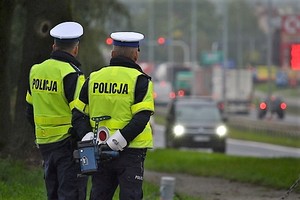 Dwóch policjantów ruchu drogowego w kamizelkach odblaskowych stojących tyłem. W tle droga i jadące pojazdy