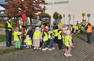 Przed budynkiem szkoły dzieci w kamizelkach odblaskowych, z transparentami, po lewej policjant