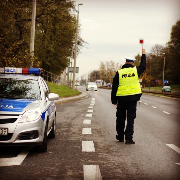 Policjant ruchu drogowego w kamizelce odblaskowej, stojący tyłem, w ręku trzyma tarcze sygnalizacyjną. Po lewej radiowóz, w tle droga i pojazdy.