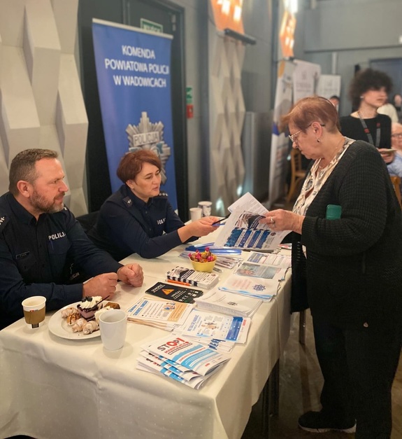 Kobieta oglądająca ulotki. Przed nią przy stoliku siedzi policjantka i policjant.
