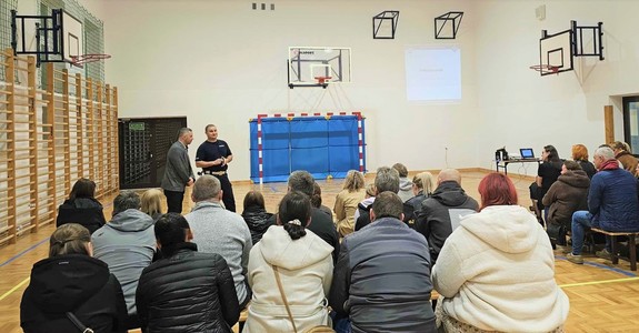Grupa osób siedzących na ławkach, są tyłem. Przed nimi policjant.