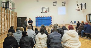 Grupa osób siedzących na ławkach, są tyłem. Przed nimi policjant.