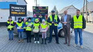 Dzieci w kamizelkach odblaskowych, policjant oraz dwóch mężczyzn, jeden ma kamizelkę odblaskową.
