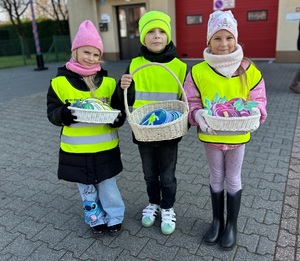 Dwie dziewczynki i chłopiec w kamizelkach odblaskowych, w rękach mają koszyczki.