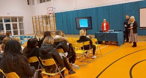 Umundurowany policjant i dwie kobiety, przed nimi grupa osób siedzących na krzesłach