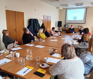 Przy stole siedzi policjant i policjanta, a wokoło osoby. W tle ekran z prezentacją.
