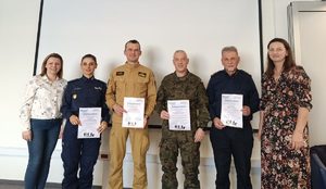 Od lewej kobieta w stroju cywilnym, policjantka, strażak, żołnierz, strażnik miejski i kobita w stroju cywilnym. Mundurowi trzymaja w rękach dyplomy.