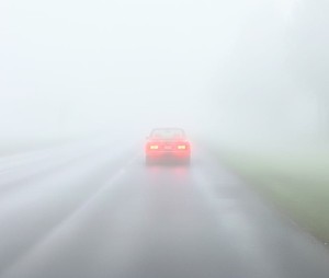 Samochód jadący po drodze podczas mgły. Ma włączone światła.