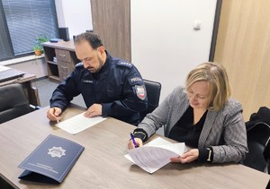 Siedzący przy stole policjant oraz kobieta w stroju cywilnym, którzy podpisują dokumenty.