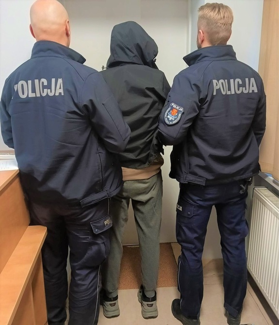 Dwóch umundurowanych policjantów, a w środku zatrzymany mężczyzna w kapturze