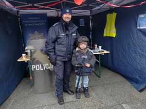 Umundurowany policjant oraz mały chłopiec ubrany w elementy policyjnego umundurowania. Stoją na tle namiotu.