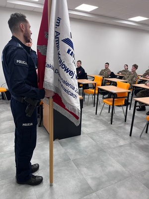 Policjant trzymający sztandar. W ławkach siedzą uczniowie w mundurach.