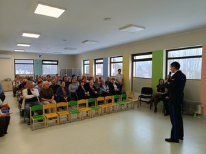 Umundurowany policjant, a przed nim osoby siedzące na krzesłach