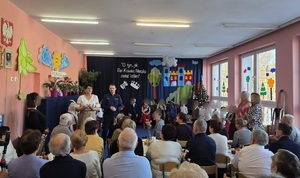 Grupa osób siedząca na krzesłach, są tyłem. Przed nimi policjant i dzieci.