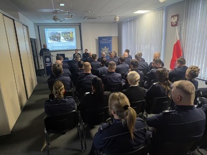 Przy mównicy stoi policjant, obok przy stoliku siedzą dwaj policjanci, a przed nimi na krzesłach siedzą policjantki i policjanci. Osoby są tyłem.