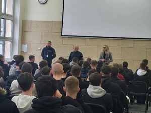 Na zdjęciu dwaj policjanci i kobieta, a przed nimi siedzący uczniowie