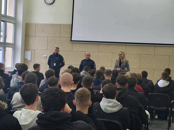 Na zdjęciu dwaj policjanci i kobieta, a przed nimi siedzący uczniowie