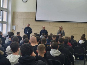 Na zdjęciu dwaj policjanci i kobieta, a przed nimi siedzący uczniowie