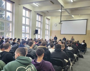 Uczniowie siedzący na krzesłach, przed nimi dwaj policjanci i kobieta w stroju cywilnym