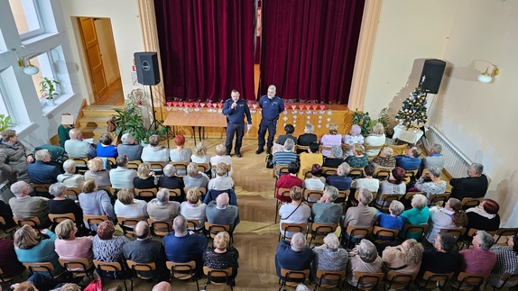 Na zdjęciu dwóch umundurowanych policjantów, a przed nimi na krzesłach siedzą osoby starsze