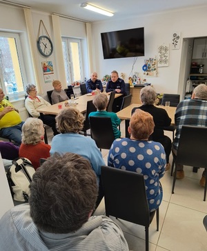 Dwóch umundurowanych policjantów siedzących przy stole, przed nimi osoby starsze siedzące na krzesłach.