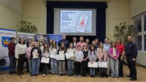 Grupa osób w tym kobiety, mężczyźni i dzieci na tle banerów i ekranu, gdzie wyświetlony jest plakat konkursu.