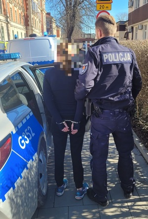 Umundurowany policjant trzymający kobietę, która ma założone kajdanki na ręce trzymane z tyłu. W tle  budynki i radiowozy.