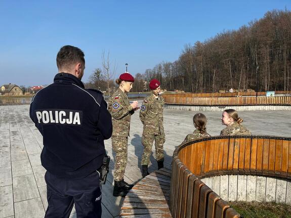 Policjant stojący tyłem i cztery dziewczyny w mundurach wojskowych, dwie z nich siedzą