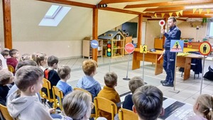 Dzieci siedzące na krzesłach, a przed nimi policjant trzymający w ręku tarcze sygnalizacyjną