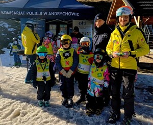 Na zdjęciu policjant oraz dwóch mężczyzn i grupa dzieci ubranych w kombinezony zimowe, kaski i gogle