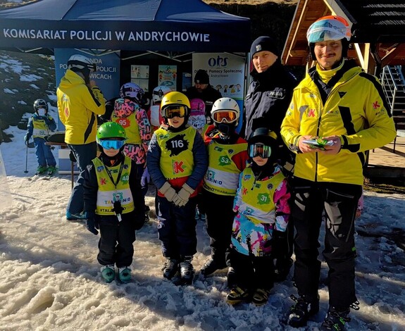 Na zdjęciu policjant oraz dwóch mężczyzn i grupa dzieci ubranych w kombinezony zimowe, kaski i gogle
