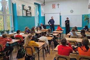 Dzieci siedzące w szkolnych ławkach przed nimi stoi kobieta i umundurowany policjant