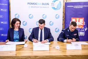 Na zdjęciu kobieta, mężczyzna oraz policjant, którzy siedzą przy stole i podpisują dokumenty