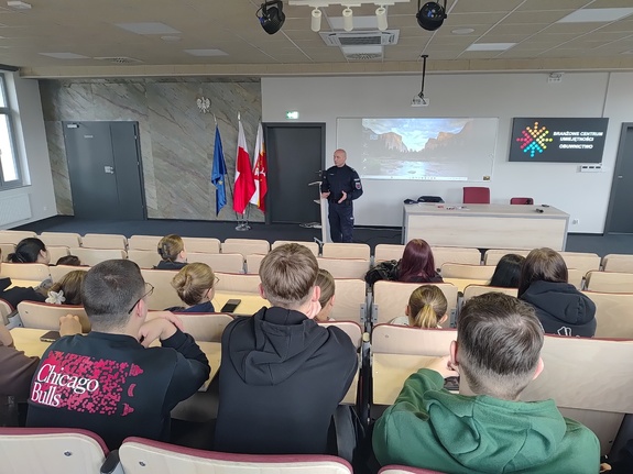 Maturzyści siedzący na auli, a przed nimi umundurowany policjant.