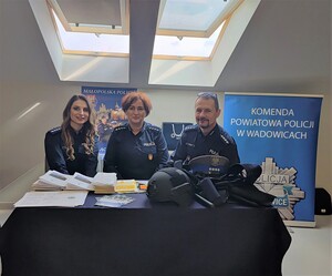 Przy stoliku na którym są ulotki i elementy umundurowania policyjnego siedzą dwie policjantki oraz policjant.. W tle baner orz plakat.