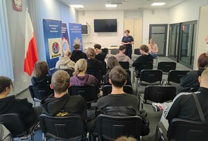 Uczniowie siedzący na krzesłach, a przed nimi stoi policjantka i na krześle siedzi policjantka w stroju cywilnym.