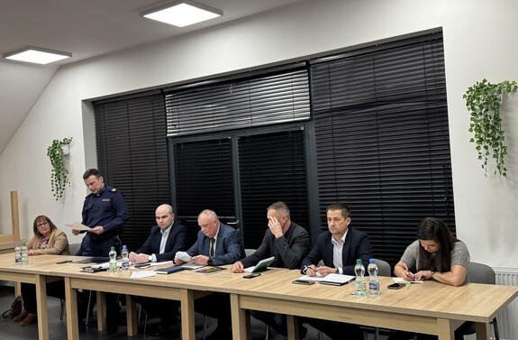 Przy stole siedzą dwie kobiety oraz czterech mężczyzn w strojach cywilnych oraz umundurowany policjant