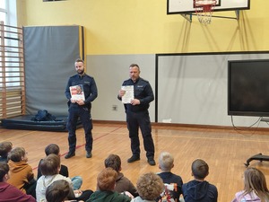 Na zdjęciu dwaj umundurowani policjanci oraz grupa uczniów siedzących przed nimi na podłodze
