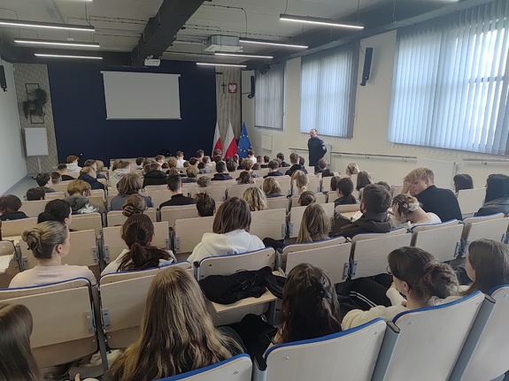 Na auli siedzą uczniowie, po prawej stronie policjant