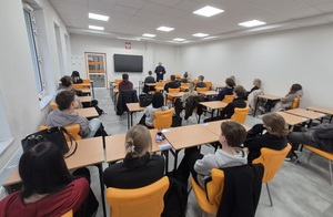 Uczniowie siedzą w szkolnych ławkach , a przed nimi policjant.