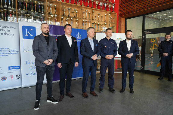 Na tle banerów czterech mężczyzn w stroju cywilnym oraz umundurowany policjant