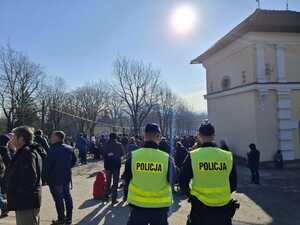 Dwaj policjanci w odblaskowych kamizelkach stojący tyłem, przed nimi tłum ludzi.