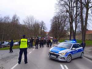 Policjant stojący tyłem, po prawej  stronie radiowóz i tłum osób