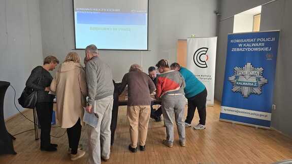 Przy stoliku siedzi policjant, a wokoło grupa seniorów zabierających ulotki.
