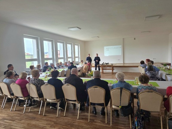 Grupa seniorów siedzących przy stolikach, przed nimi dwaj umundurowani policjanci.